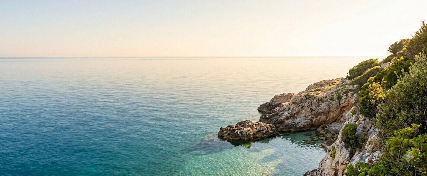 Tranquil Mediterranean coastline featuring clear turquoise water, rocky cliffs covered in lush green vegetation, and bright sunlight setting a summer vibe for field research stories.